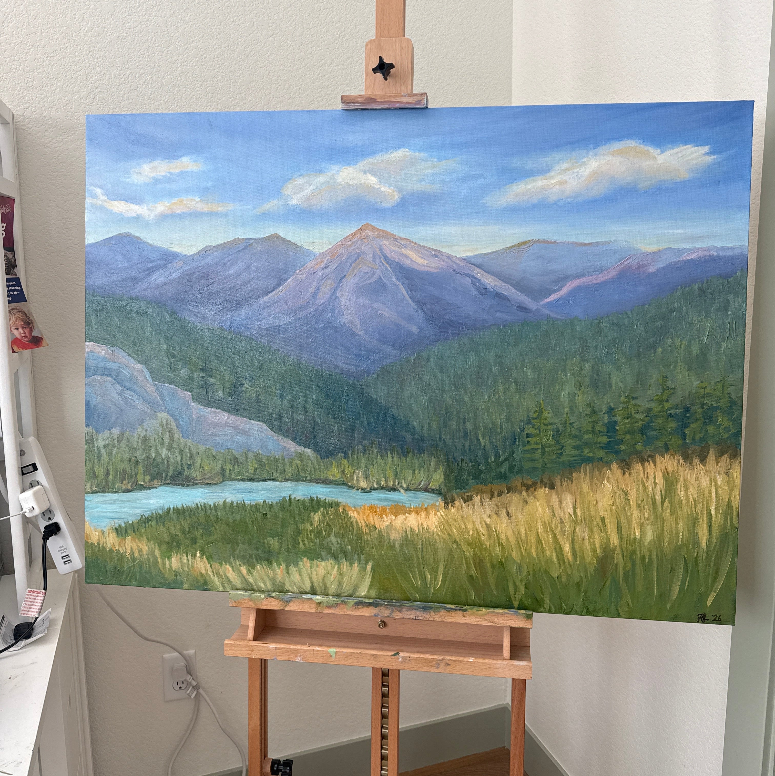 Painting of a landscape with blue sky and green mountains on an easel.